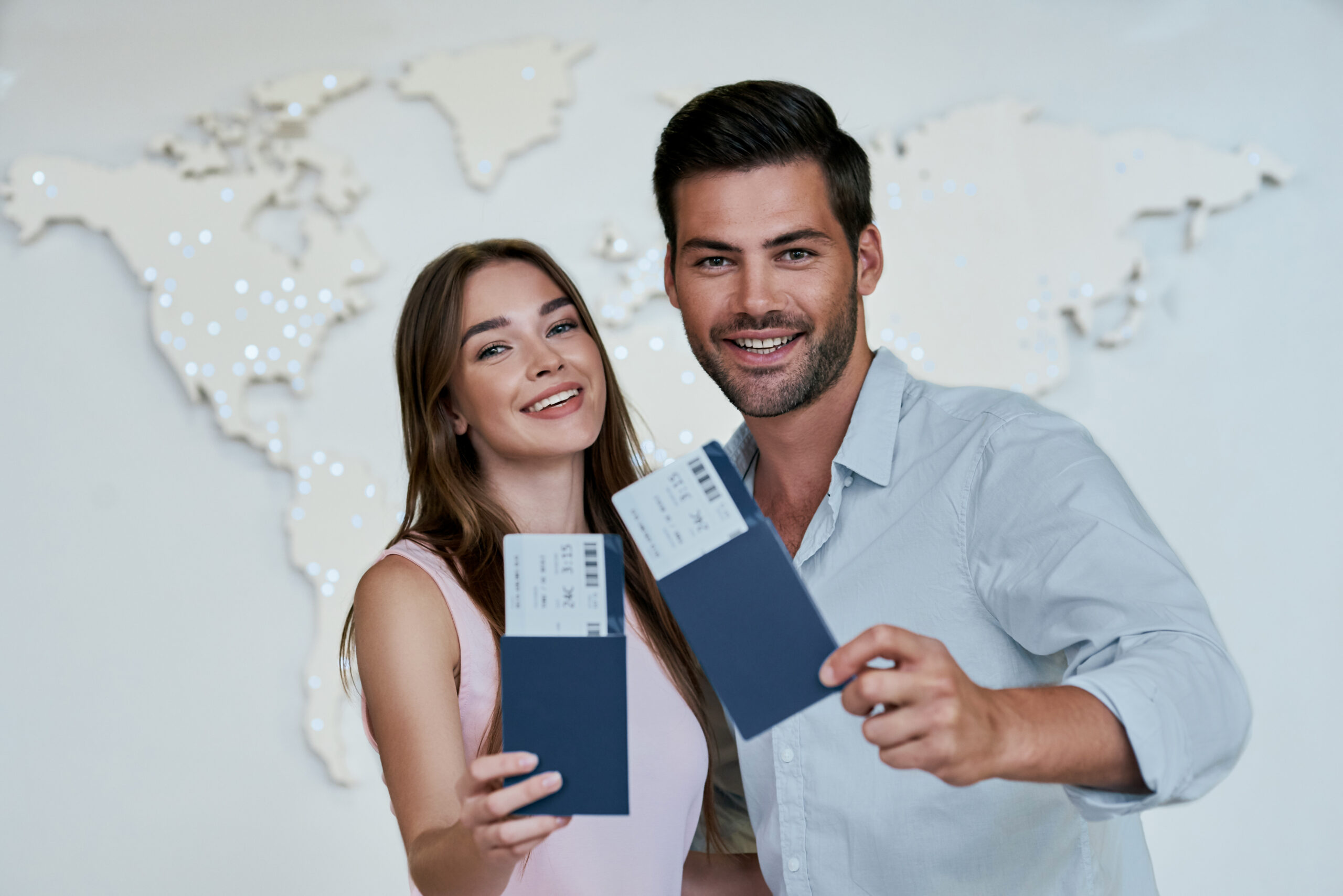 Portrait of joyful glad couple holding passport with flying tickets in hands pointing with forefinger looking at camera on map background